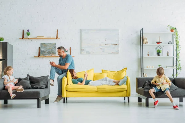 Mutter und Vater mit zwei Kindern nutzen Smartphones, während sie zu Hause auf dem Sofa liegen — Stockfoto