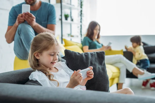 Selektiver Fokus des niedlichen Kindes mit Smartphone in der Nähe von Vater, Mutter und Bruder mit Smartphone zu Hause — Stockfoto