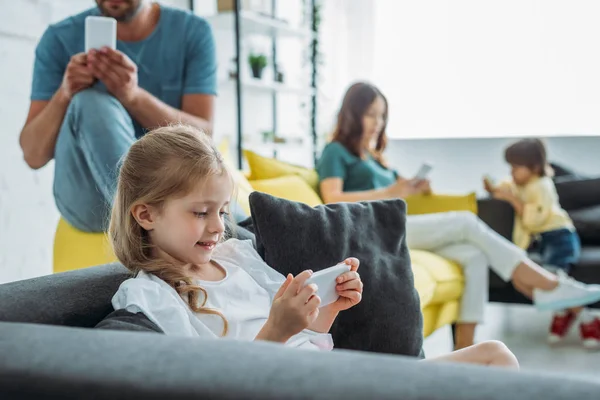 Messa a fuoco selettiva del bambino carino utilizzando smartphone vicino a padre, madre e fratello utilizzando smartphone a casa — Foto stock