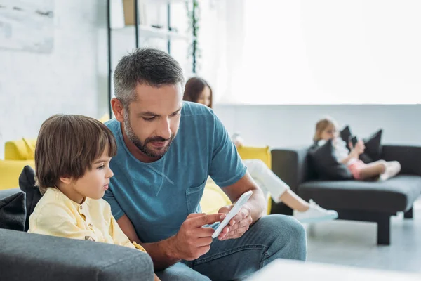 Messa a fuoco selettiva del padre mostrando smartphone al figlio carino vicino a madre e figlia utilizzando smartphone a casa — Foto stock