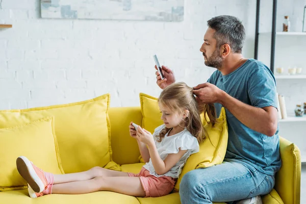 Padre tocando el pelo hijas mientras está sentado en el sofá y el uso de teléfonos inteligentes juntos - foto de stock