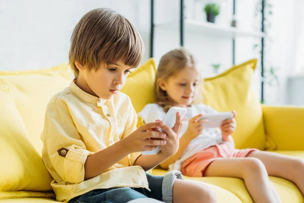 Selective focus of cute boy with sister sitting on sofa and using smartphones at home — Stock Photo