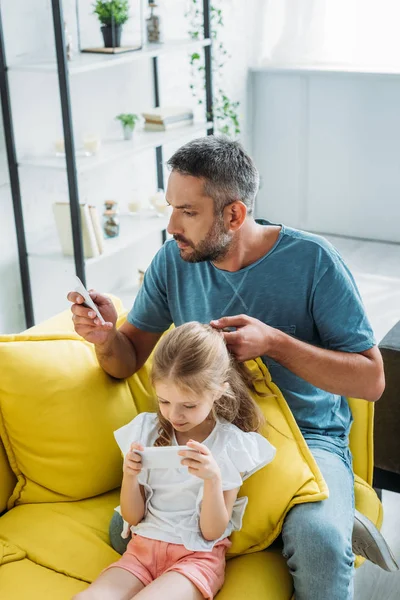 Concentrated father touching hair of daughter while sitting on sofa and using smartphones together — Stock Photo