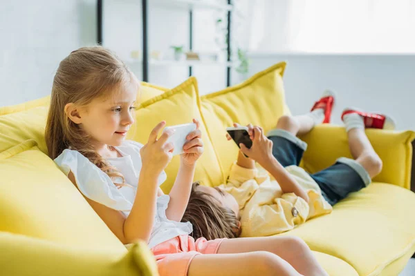 Очаровательный ребенок сидит со смартфоном рядом брат лежит на диване и с помощью смартфона дома — стоковое фото