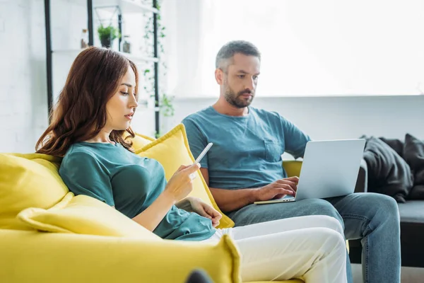 Attraktive Frau mit Smartphone sitzt neben Ehemann und benutzt Laptop zu Hause — Stockfoto