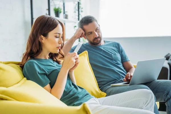 Hombre reflexivo con el ordenador portátil sentado en el sofá cerca de la esposa utilizando el teléfono inteligente en casa - foto de stock