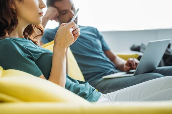 Vue partielle de la femme en utilisant un smartphone tout en étant assis près du mari en utilisant un ordinateur portable — Photo de stock
