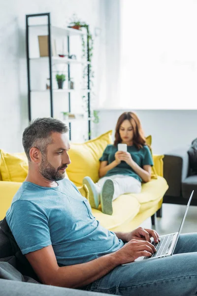 Enfoque selectivo del hombre concentrado usando el ordenador portátil mientras está sentado en el sofá cerca de la esposa utilizando el teléfono inteligente - foto de stock
