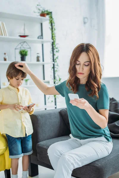 Woman with smartphone touching head of son standing near and using smartphone — Stock Photo