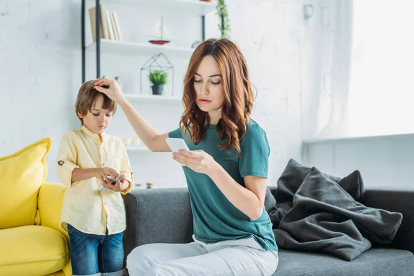 Bonita mujer con teléfono inteligente tocando la cabeza de lindo hijo de pie cerca y utilizando el teléfono inteligente - foto de stock