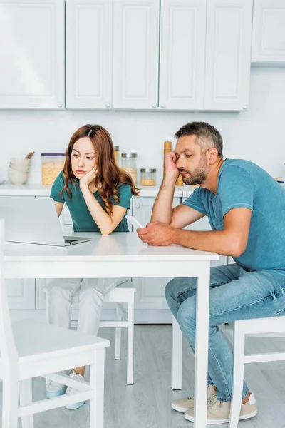 Nachdenkliche Frau nutzt Laptop, während sie am Küchentisch neben Ehemann mit Smartphone sitzt — Stockfoto