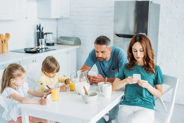 Familia usando teléfonos inteligentes mientras está sentado en la mesa de la cocina cerca del desayuno servido - foto de stock