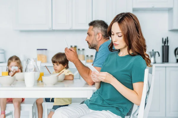 Esposa e marido com smartphones sentados na mesa da cozinha com café da manhã servido perto de crianças usando smartphones — Fotografia de Stock