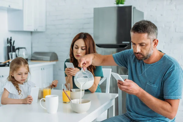 Мужчина наливает молоко в миску, сидя за кухонным столом с подаваемым завтраком рядом с женой и дочерью, используя смартфоны — стоковое фото
