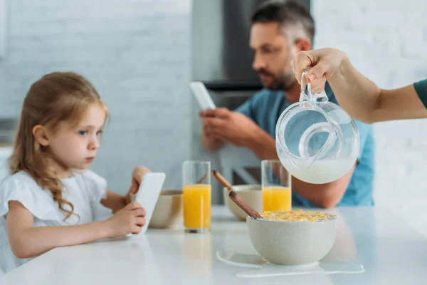 Vista cortada de mulher derramando leite em tigela com flocos perto de filha e marido usando smartphones — Fotografia de Stock