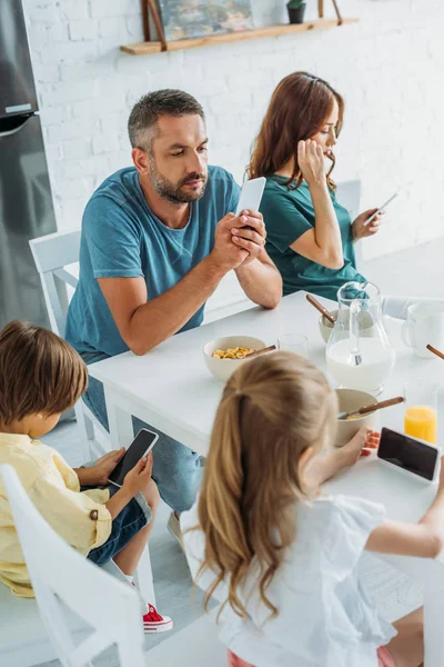 Familia sentada en la mesa de la cocina cerca del desayuno servido y utilizando teléfonos inteligentes — Stock Photo