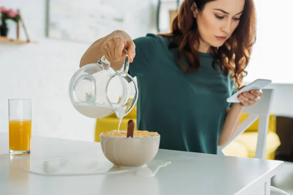 Frau überfüllt Schüssel mit Flocken mit Wasser, während sie am Küchentisch sitzt und Smartphone benutzt — Stockfoto