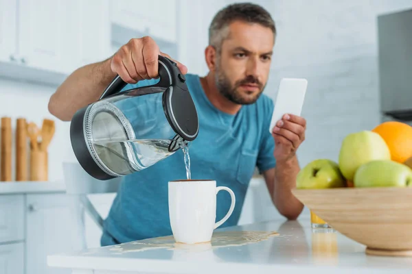 Selektiver Fokus des Mannes, der Tasse mit Wasser überfüllt, während er am Küchentisch sitzt und Smartphone benutzt — Stockfoto