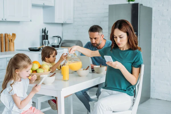 Mujer con teléfono inteligente verter jugo de naranja en vidrio mientras está sentado cerca de la familia usando teléfonos inteligentes en la cocina — Stock Photo