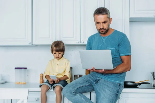 Father using laptop and and son using smartphone while sitting on kitchen counter together — Stock Photo