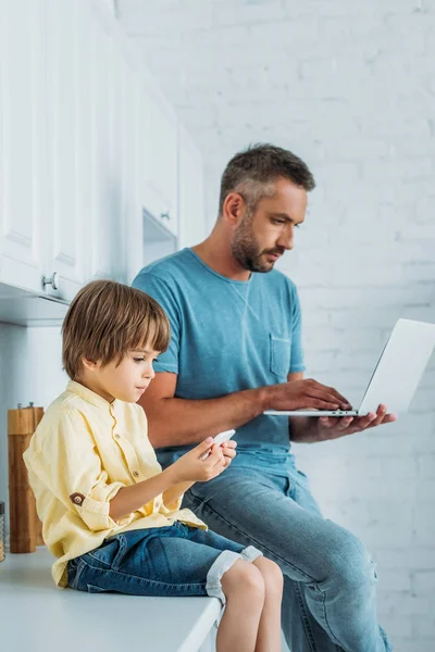 Мила дитина використовує смартфон, сидячи на кухонній стійці біля батька, використовуючи ноутбук — стокове фото