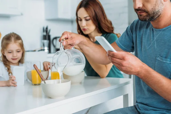 Hombre con teléfono inteligente verter leche en un tazón mientras está sentado en la mesa de la cocina cerca de la esposa y la hija usando teléfonos inteligentes - foto de stock
