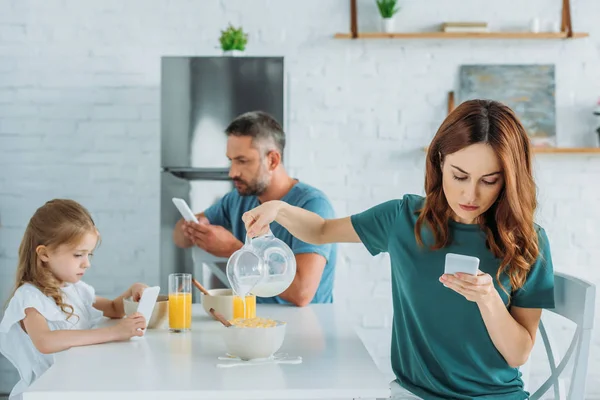 Frau mit Smartphone überfüllt Schüssel mit Milch, während sie am Küchentisch in der Nähe von Mann und Tochter mit Smartphones sitzt — Stockfoto