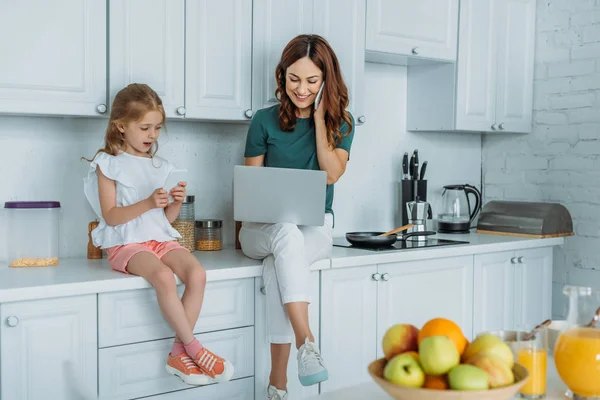 Woman talking on smartphone and using laptop while sitting on kitchen counter near daughter using smartphone — Stock Photo