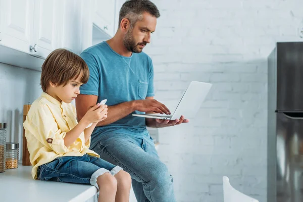 Hombre usando el ordenador portátil mientras está sentado en el mostrador de la cocina cerca de hijo usando smarthone - foto de stock