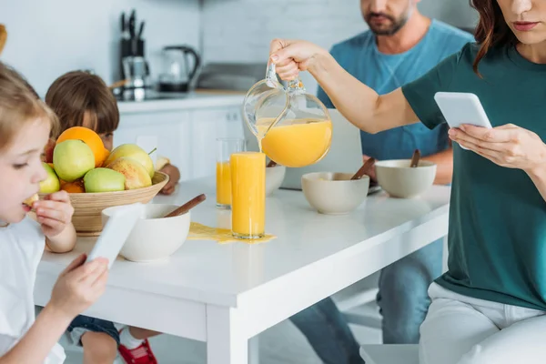 Vista recortada de la mujer con teléfono inteligente vaso de relleno con jugo de naranja mientras está sentado en la mesa de la cocina cerca de la familia usando smartpones - foto de stock