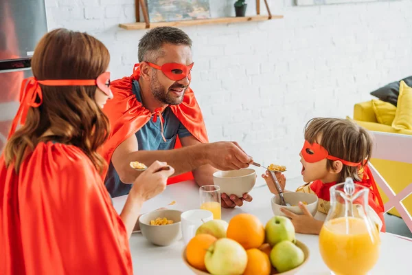 Cheerful family in superheroes costumes having breakfast in kitchen together — Stock Photo