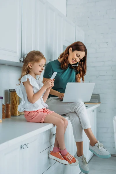 Улыбающаяся женщина разговаривает на смартфоне и с помощью ноутбука, сидя на кухонном столе рядом с дочерью со смартфоном — стоковое фото