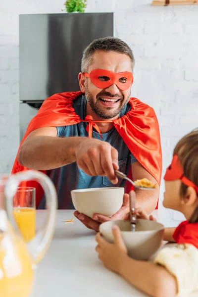 Pai e filho alegres em fantasias de super-heróis tomando café da manhã na cozinha juntos — Fotografia de Stock