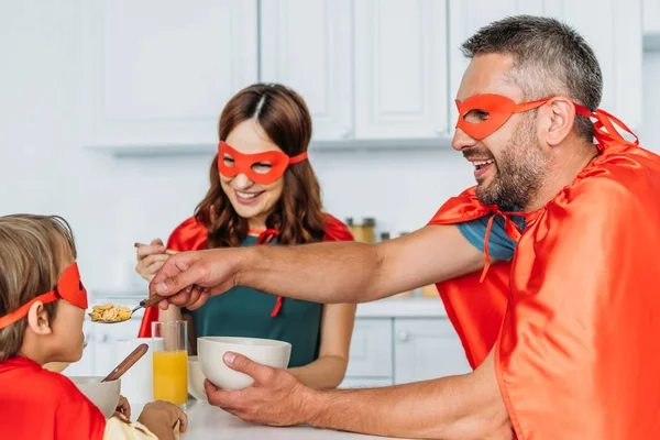 Família em trajes de super-heróis tomando café da manhã enquanto o pai alimentando o filho com flocos — Fotografia de Stock