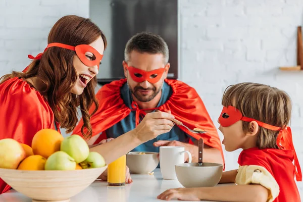 Happy mother feeding son with flakes while sitting at kitchen table in superheroes costumes — Stock Photo