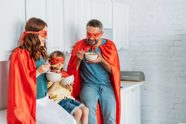 Glückliche Eltern mit Sohn in Superheldenkostümen beim Frühstück, während er auf der Küchentheke sitzt — Stockfoto