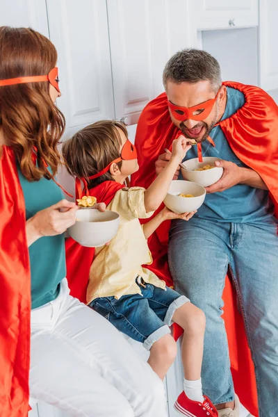 Веселая семья в костюмах супергероев ест хлопья из миски, сидя на кухонном столе — стоковое фото