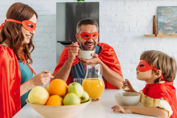 Pai feliz, mãe e filho em trajes de super-heróis tomando café da manhã juntos — Fotografia de Stock