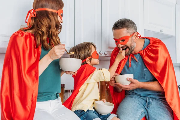 Happy family in costumes of superheroes eating breakfast while son feeding father with flakes — Stock Photo