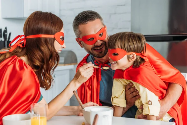 Madre alimentación hijo con escamas, mientras que la familia en trajes de superhéroes desayunando - foto de stock