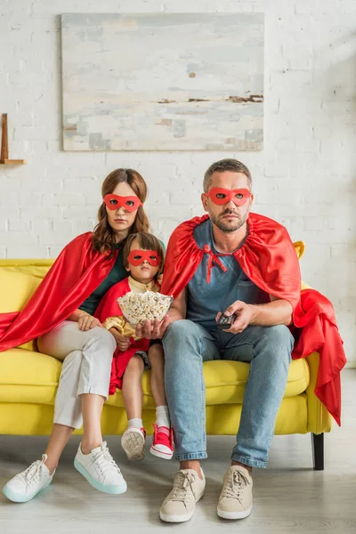 Família em trajes de super-heróis sentados no sofá amarelo com pipocas e assistindo tv — Fotografia de Stock