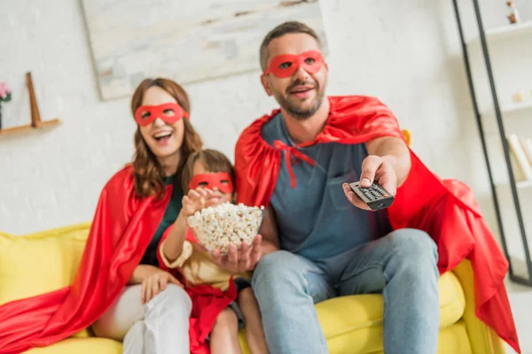 Selektiver Fokus des Vaters mit Fernbedienung, während die Familie in Superheldenkostümen auf dem Sofa mit Popcorn sitzt und fernsieht — Stockfoto