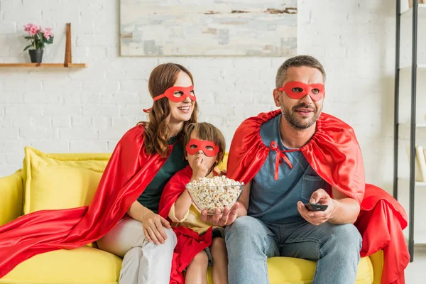 Familia alegre en trajes de superhéroes sentados en el sofá y viendo la televisión - foto de stock