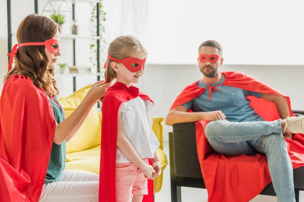 Mother in costume of superhero dressing daughter while father sitting on sofa — Stock Photo