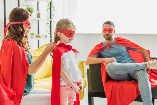 Madre disfrazada de superhéroe vestidor hija mientras el padre sentado en el sofá - foto de stock