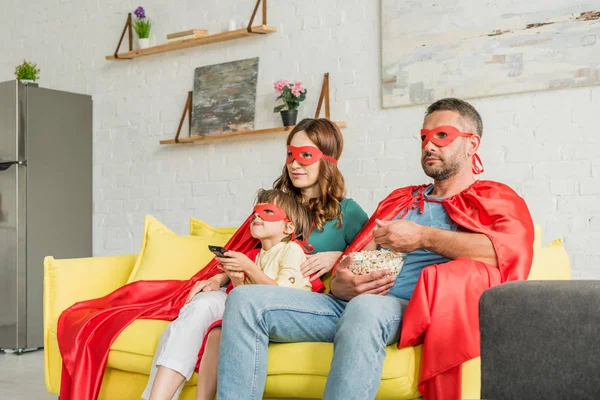 Family in costumes of superheroes sitting on sofa, eating popcorn and watching tv — Stock Photo