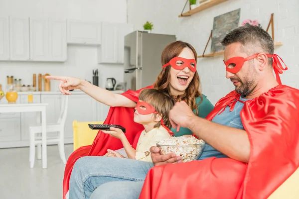 Cheerful mother pointing with finger while family in costumes of superheroes sitting on sofa and watching tv — Stock Photo