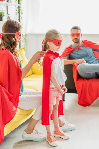 Enfoque selectivo de la madre vestir hija en traje de superhéroe, mientras que el padre sentado en el sofá - foto de stock