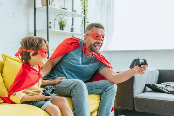 KYIV, UCRANIA - 5 de julio de 2019: Feliz padre e hijo con disfraces de superhéroes sentados en el sofá y jugando videojuegos - foto de stock