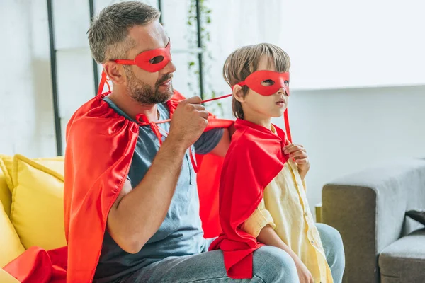 Padre en traje de superhéroe poniéndose máscara roja en hijo - foto de stock